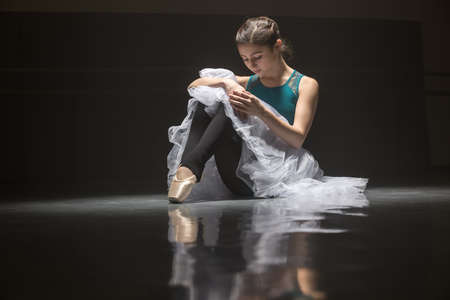 Cute ballerina sits on the floor in the dance hall and looks at her hands. She has brown hair. She...