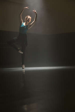 Young ballet dancer stands with raised arms on a pointe shoe in the dark dance hall. Narrow light falls down on her. Low key photo.の写真素材