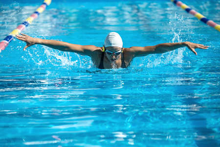 Energetic female swimmer swims in the butterfly style in the swimming pool. Woman wears a black-lime swimsuit, a white swim cap and swim glasses. Outdoors. Horizontal.の写真素材