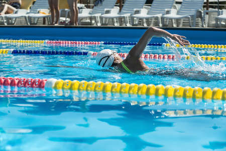 Backside view of the female swimmer who swims in the swimming pool outdoors. She wears a black-lime swimsuit, a white swim cap and swim glasses. Horizontal.の写真素材