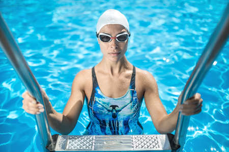Nice sportive girl stands on the pool ladder in the swim pool and holds hands on it. She wears a blue-black swimsuit, a white swim cap and swim glasses. Woman looks into the camera with a smile.の写真素材
