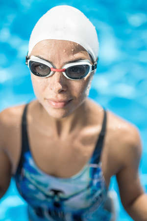 Athletic wet girl in the swimming pool. She wears a blue-black swimsuit, a white swim cap and swim glasses. Woman looks forward with a smile. Close-up photo. Vertical.の写真素材