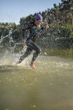 Energetic female runner runs on the water outdoors. She wears a dark swimrun suit, violet cap, swim glasses and a stopwatch. Water splashes are around her body. Sun shines at her body. Vertical.の写真素材