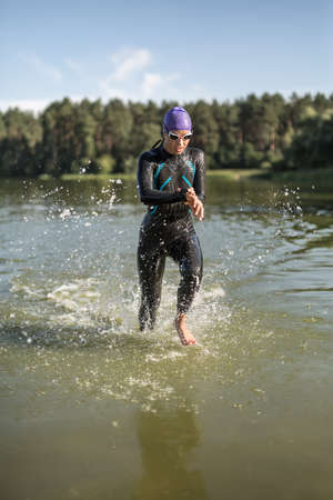 Active female runner runs on the water outdoors. She wears a dark swimrun suit, violet cap, swim glasses. Girl looks at stopwatch. Water splashes are around her body. Sun shines at her body. Vertical.の写真素材