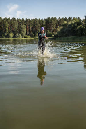 Sportive female runner runs on the water outdoors. She wears dark swimrun suit, violet cap, swim glasses. Girl looks at the stopwatch. Water splashes are around her body. Girl reflected in the water.の写真素材