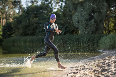 Charming female runner runs on the waterfront outdoors. She wears dark swimrun suit, violet cap, swim glasses. Girl looks at the stopwatch. Water splashes are around her body. Sun shines at her body.の写真素材
