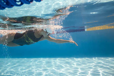 Sportive female swimmer swims underwater in the swimming pool outdoors. She wears a black-gray swimsuit with patterns, a white swim cap and swim glasses. Sunlight falls from above. Horizontal.の写真素材