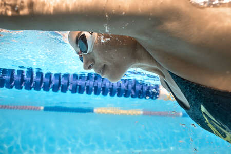 Incredible close-up photo of woman who swims underwater in the swimming pool outdoors. She wears a gray swimsuit, a white swim cap and white swim glasses. Sunlight falls from above. Horizontal.の写真素材
