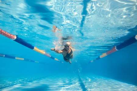 Motion photo of a girl who swims underwater in the swim pool outdoors. She wears a black-gray swimsuit with patterns, a white swim cap and swim glasses. Sunlight falls from above. Horizontal.の写真素材