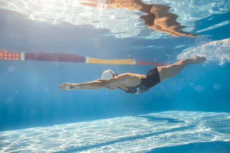 Great swimmer swims in crawl style underwater in the swim pool outdoors. She wears black-gray swimsuit, white swim cap and swim glasses. Sunlight falls from above. Her body reflected in water surface.の写真素材