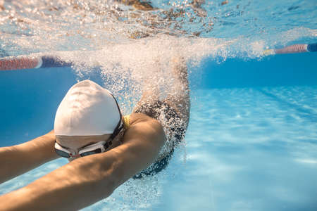 Emotional close-up photo of woman who swims underwater in the swimming pool outdoors. She wears dark swimsuit, white swim cap and swim glasses. Sunlight falls from above. Splashes are around her body.の写真素材