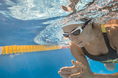 Cool close-up photo of woman who swims underwater in the swimming pool outdoors. She wears a gray-lime swimsuit, a white swim cap and white swim glasses. Sunlight falls from above. Horizontal.の写真素材