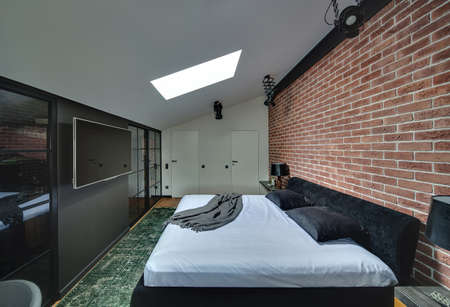 Contemporary bedroom with brick wall and parquet with green carpet on the floor. There is bed with pillows, racks with lamps and decorations, black wall with cupboards and TV, white wall with doors.の写真素材