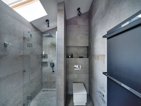 Bathroom in modern style with gray textured tiles on the floor and walls. There is shower with glass door, white toilet, black panel with two chrome towel rails. At the top there are lamps and window.の写真素材