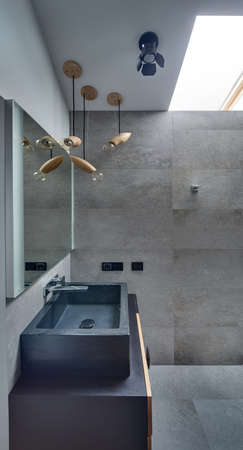 Modern bathroom with gray textured tiles on the floor and walls. There is sink with tap and mirror. Under the sink there is locker. At the top there are different lamps and window. Vertical.の写真素材
