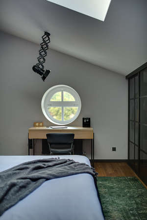 Round window at the white wall. Under it there is table with laptop and clock and black chair. Also there is bed with coverlet, black wardrobe, lamp and window at the top, green carpet on the floor.の写真素材