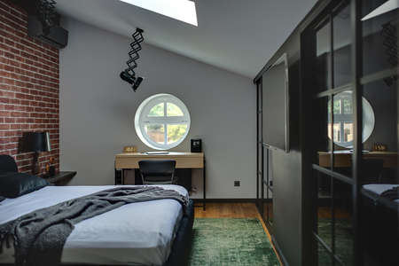 Amazing modern bedroom with brick wall and parquet with green carpet. There is bed, rack with lamp, round window, table with chair, wall with TV and wardrobes. At the top there are lamps and window.の写真素材