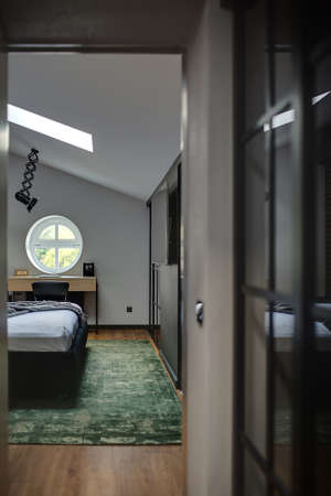 Pretty modern bedroom with light walls and parquet with green carpet on the floor. There is bed, round window, table with chair, wall with TV and wardrobes. At the top there is black lamp and window.の写真素材