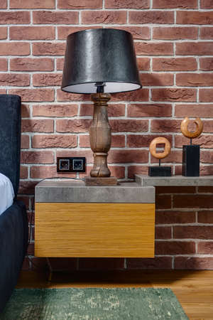 Wooden lamp with black lampshade on the rack on the brick wall background. On the right there is rack with decorations. On the left there is bed. On the floor there is parquet and green carpet.の写真素材