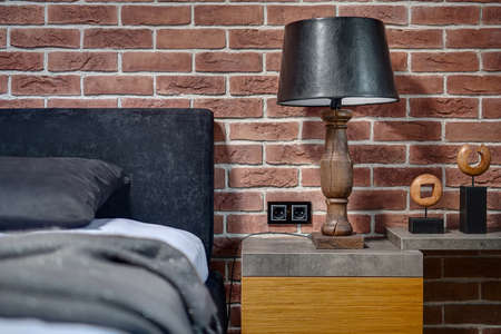 Old style wooden lamp with black lampshade on the rack on the brick wall background. On the right there is rack with decorations. On the left there is bed with pillow and coverlet. Horizontal.の写真素材