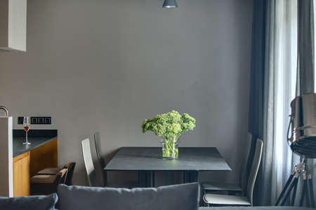Dark table with vase with green flowers and four chairs on the gray wall background. On the right there is window with curtains. On the left there is kitchen island. At the top there is lamp.の写真素材