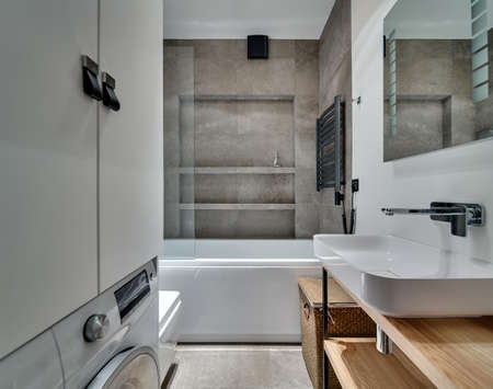 Bathroom in modern style with textured tiles, white sink with tap and mirror, white bath. Opposite the sink there is white locker and washing machine under it. Under the sink there are wooden racks.の写真素材
