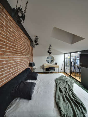 Trendy modern bedroom with cloakroom with glowing lamps, brick wall, bed, table with chair, TV, round window on the wall and window at the top. On the floor there is parquet and green carpet.の写真素材