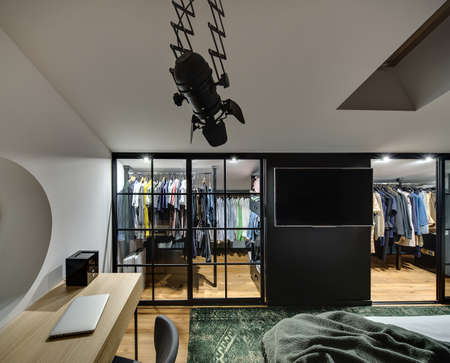 Elegant modern bedroom with cloakroom with glowing lamps, white walls. There is bed, table with laptop and chair, TV, window at the top. There are a lot of clothes in the cloakroom. Horizontal.の写真素材