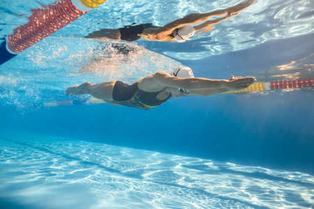 Women swim in the swim pool outdoors. One girl swims in crawl style underwater. She wears a swimsuit, a swim cap and swim glasses. Her body reflected in water surface. Sunlight falls from above.の写真素材