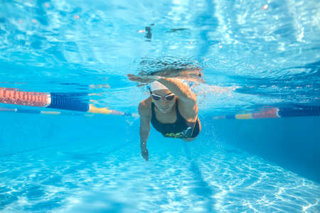 Attractive girl swims underwater in the swimming pool with other people outdoors. She wears a black-gray swimsuit with patterns, a white swim cap and swim glasses. Sunlight falls from above.の写真素材