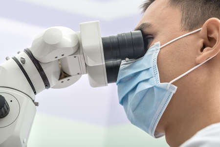 Young dentist looks into the dental microscope in the clinic on the blurry wall background. He wears a white uniform and a blue medical mask. Horizontal.の写真素材