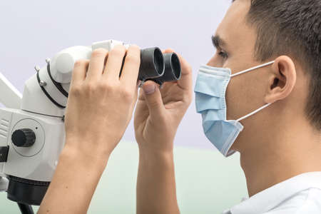 Doctor looks into the dental microscope which he holds with his hands in the clinic on the wall background. He wears a white uniform and a blue medical mask. Horizontal.の写真素材