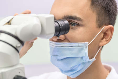 Image of a doctor who looks into the dental microscope on the light background. He wears a blue medical mask. Close-up. Horizontal.の写真素材