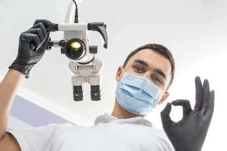 Happy dentist stands in the clinic and shows OK symbol. Man holds a glowing dental microscope with right hand. He wears a white uniform, black gloves and a blue medical mask. View from the bottom.の写真素材