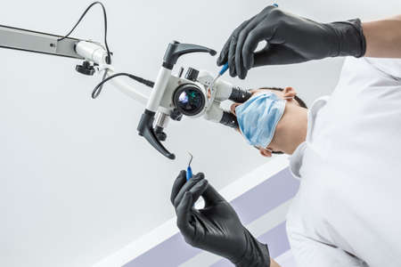 Male doctor is using the glowing dental microscope in the clinic. He is wearing a white uniform, black gloves and a blue medical mask. Man holds a dental bur and a mirror. View from the bottom.の写真素材