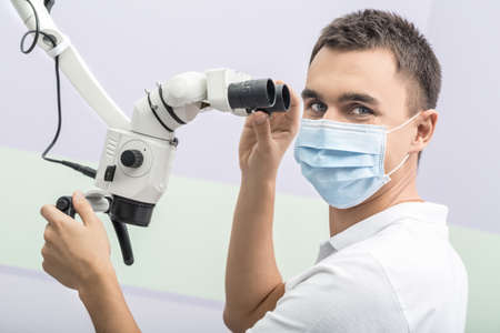 Cheerful doctor is looking into the camera while holding microscope with his hands in the clinic on the wall background. He wears a white uniform and a blue medical mask. Horizontal.の写真素材