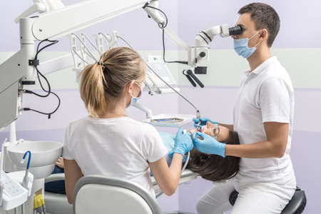 Patient in goggles on a dental chair and a dentist with assistant who sit next to her. Man looks on her teeth using a dental microscope and holds dental instruments. Woman holds an air water syringe.の写真素材