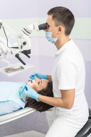 Nice girl with patient bib on a dental chair and dentist who sits next to her. He looks on her teeth using dental microscope and holds dental bur and mirror. Vertical.の写真素材
