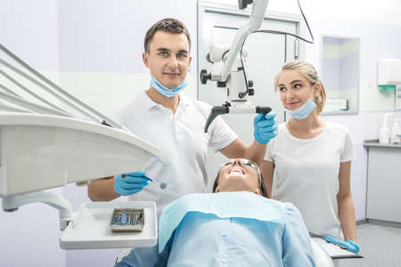 Pretty dentist and an assistant stand next to girl on a patient chair in a dentist's office. Doctor holds a dental microscope and a dental mirror. They wears white uniform with blue masks and gloves.の写真素材