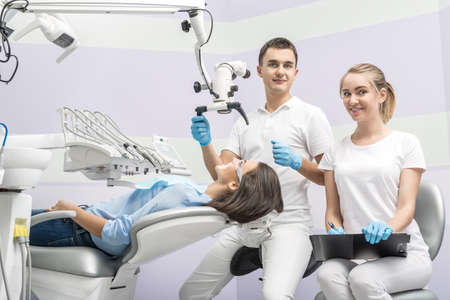 Dentist and a smiling assistant sit next to girl on a patient chair in a dentist's office. Doctor holds a dental microscope. Blonde holds a pen and a paper clipboard. They both wears white uniform.の写真素材