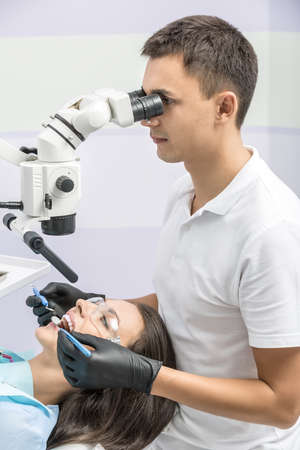 Nice dentist sits on a chair and looks at the girl's teeth using a dental microscope. He holds a dental bur and mirror. Doctor wears a white uniform and black gloves. Woman has protective glasses.の写真素材
