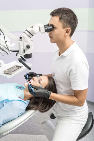 Friendly dentist sits on a chair and looks at the girl's teeth using a dental microscope. He holds a dental bur and mirror. Doctor wears a white uniform and black gloves. Woman has protective glasses.の写真素材
