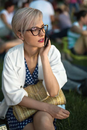 Blonde girl in glasses talks on a cell phone on the blurry background outdoors. She sits on the...
