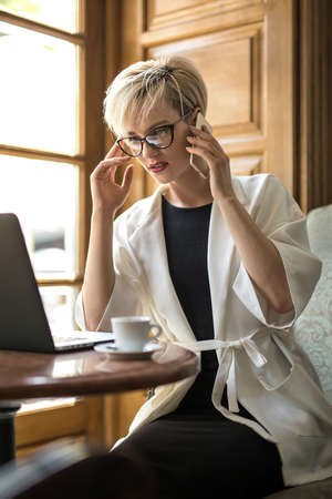 Cute blonde girl in glasses sits on armchair at the table in the restaurant and talks on cell phone. Her right hand is on the head, she looks at the laptop. Woman wears black dress and a white cloak.の写真素材