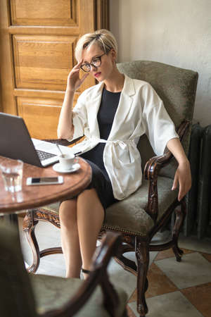Young blonde girl in glasses sits on the armchair at the table in the restaurant and looks at the laptop. She holds the right hand on her head. Woman wears black dress and a white cloak. Vertical.の写真素材