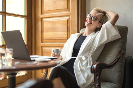 Dreamy blonde girl in glasses sits on the armchair at the table with a laptop in the cafe. She leans on armchair with a smile and holds a cup in right hand. Woman wears black dress and a white cloak.の写真素材