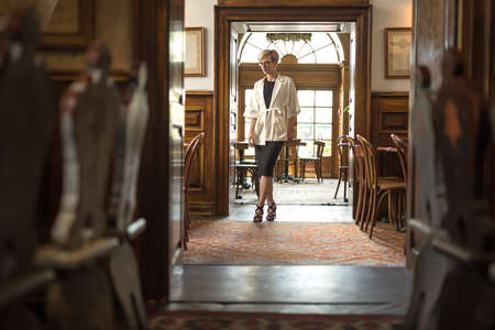 Beautiful blonde girl in the glasses stands near the doorway in the hall of the restaurant. Her legs are crossed, hands are along the body. Woman wears a black dress and sandals, a white coat.の写真素材