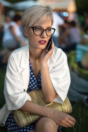 Pretty blonde girl in glasses talks on a cell phone on the blurry background outdoors. She sits on the grass in a blue dress with white pateerns and a white cloak. Woman holds a bag. Vertical.の写真素材