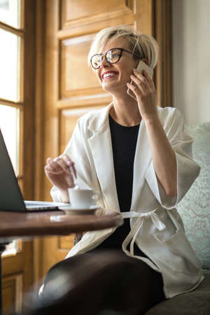 Joyous blonde girl in glasses sits on the armchair at the table with laptop in the cafe and talks on the cell phone. She puts the spoon in the white cup. Woman wears black dress and a white cloak.の写真素材