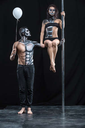 Strong couple of pole dancers in the dark studio. Man holds a pylon with the left hand and holds a balloon in the right. Girl sits on the guy's left arm. They have a horrific body-art. Vertical.の写真素材